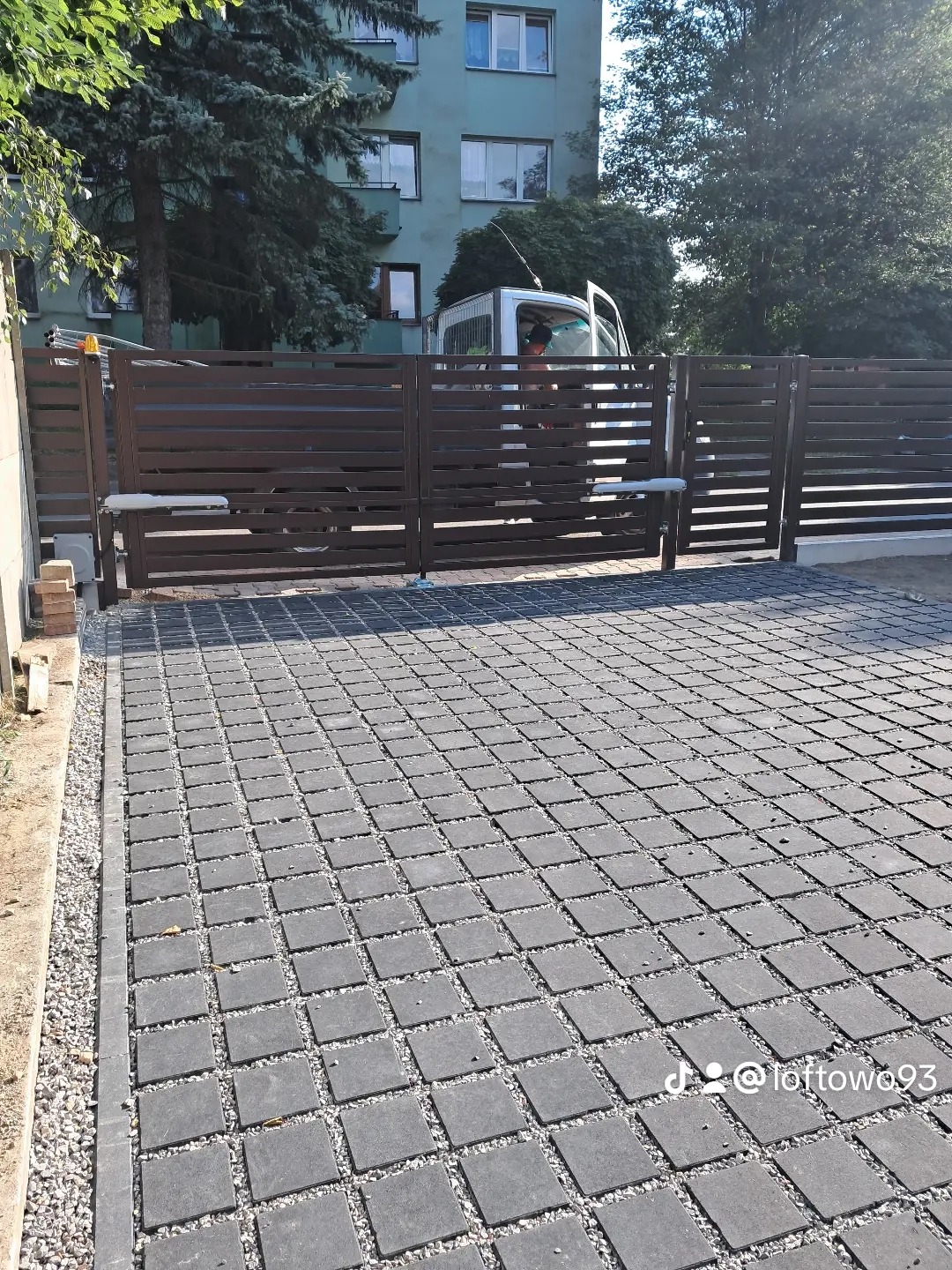 Nowoczesna, brązowa brama przesuwna z poziomych profili, z automatyką, na tle bloku mieszkalnego i zaparkowanego busa. Kostka brukowa na podjeździe.