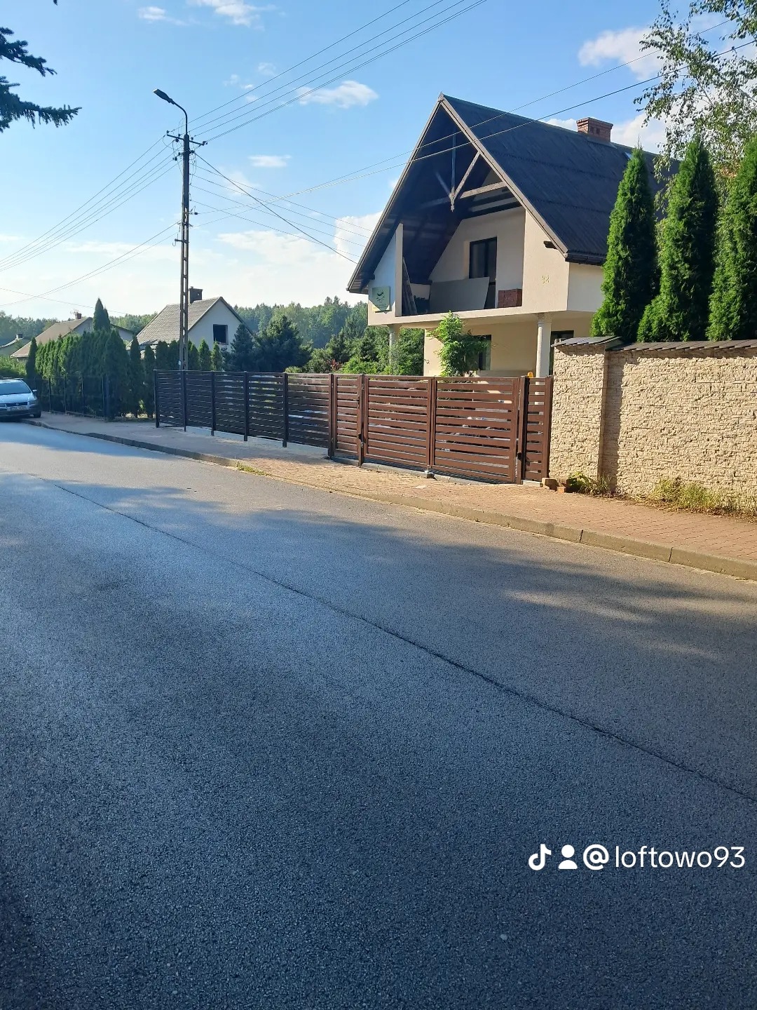 Nowoczesne ogrodzenie panelowe z poziomych desek w kolorze brązowym, oddzielające posesję od ulicy. W tle dom z balkonem i zielone tuje.