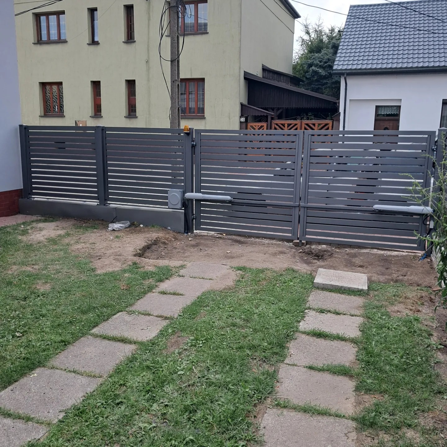Nowoczesna, pozioma brama metalowa w kolorze grafitowym z automatyką, widoczna z perspektywy ścieżki z płyt betonowych na tle budynków mieszkalnych.
