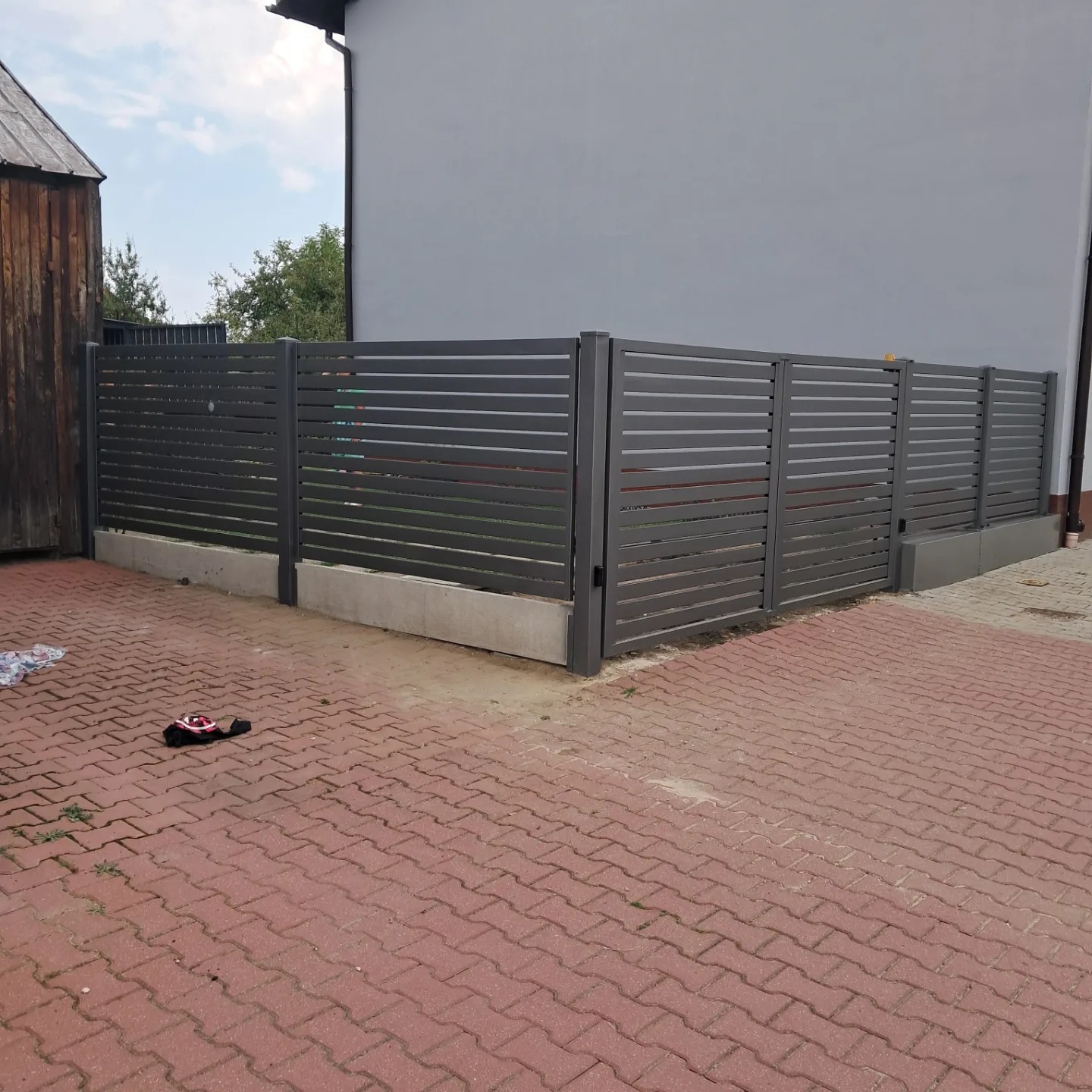 Nowoczesne, poziome ogrodzenie metalowe w kolorze grafitowym, z betonową podmurówką, na tle szarego budynku i czerwonej kostki brukowej. Na pierwszym planie zabawka.
