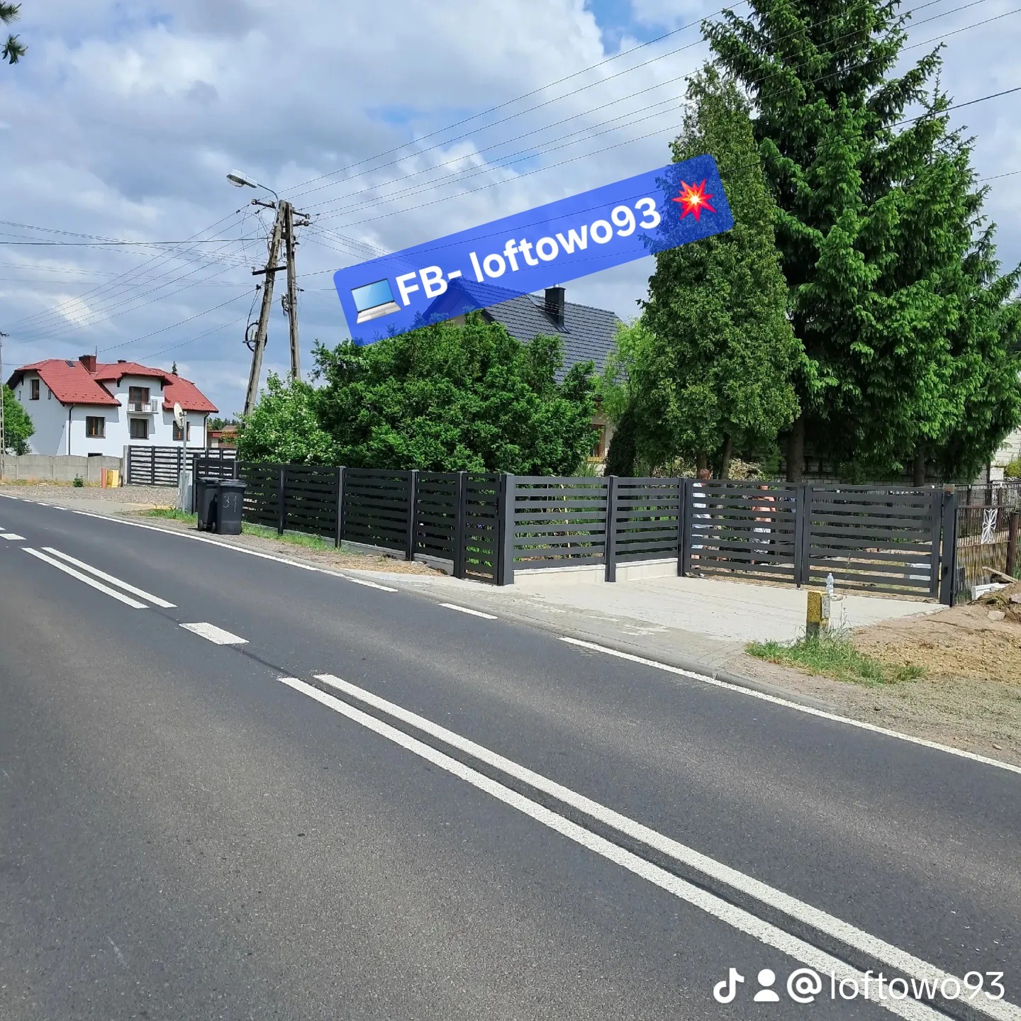 Nowoczesne, poziome ogrodzenie metalowe w kolorze antracytowym, widoczne z perspektywy ulicy. Brama wjazdowa otwarta, zieleń w tle, domy po obu stronach.