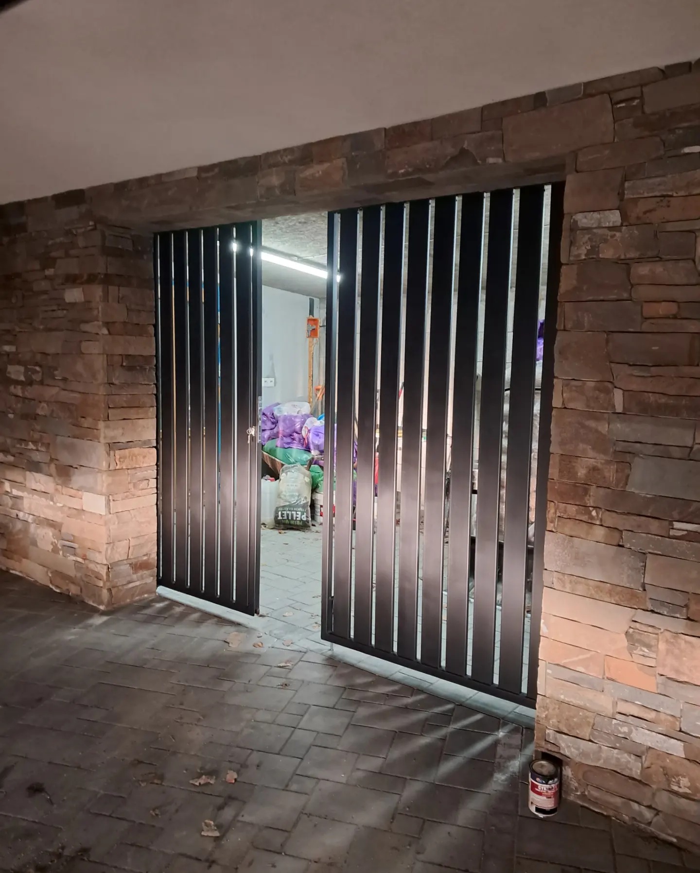 Nowoczesna, czarna brama lamelowa, częściowo otwarta, w kamiennym murze. Widoczny fragment podjazdu z kostki brukowej i wnętrze garażu z workami.