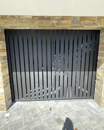 Nowoczesna, grafitowa brama wjazdowa z pionowych profili, osadzona w kamiennym murze. Widoczne detale kostki brukowej i fragment elewacji.