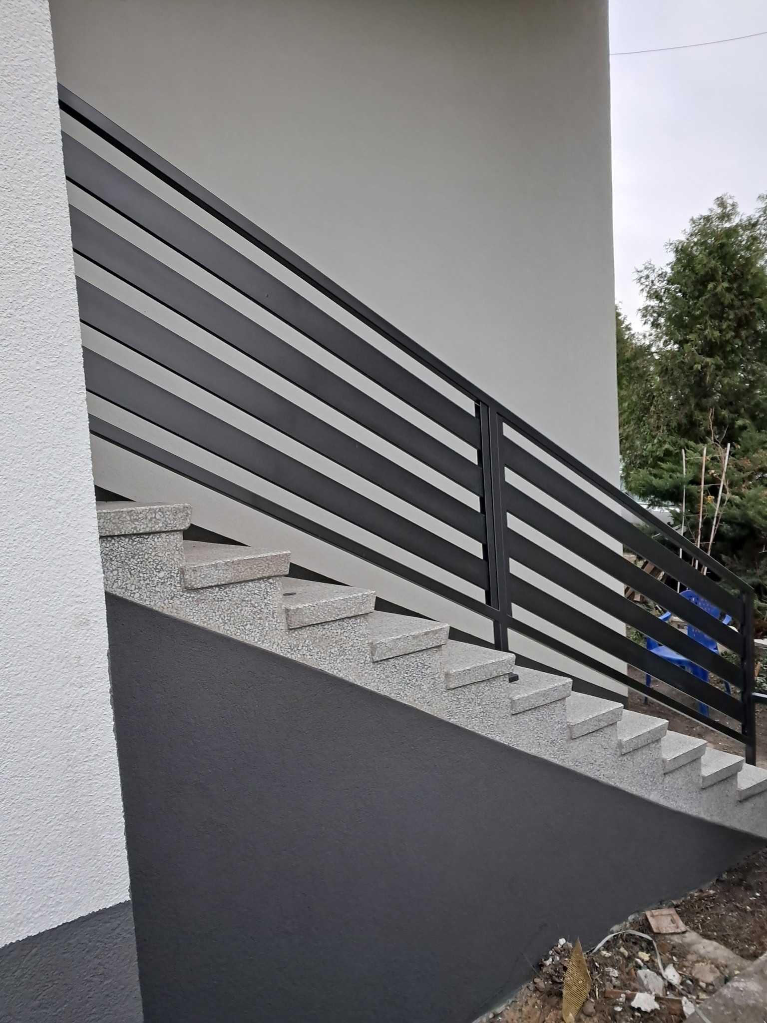 Zewnętrzne schody z szarego kamienia z nowoczesną, poziomą balustradą z czarnych, metalowych profili. Minimalistyczny design, jasne tło budynku.