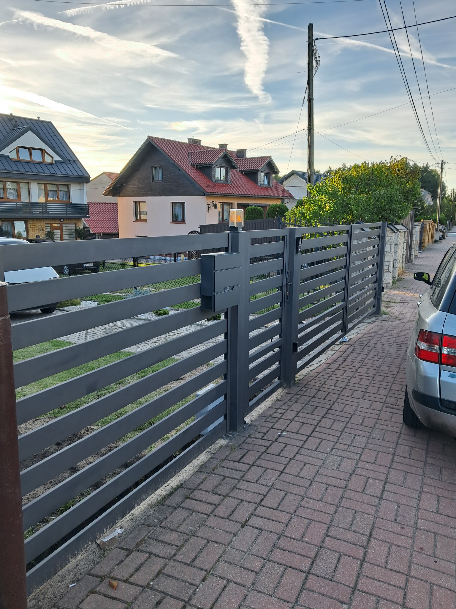 Nowoczesne, poziome ogrodzenie metalowe w kolorze grafitowym, zintegrowane ze skrzynką na listy. Widok z zewnątrz posesji, kostka brukowa, domy w tle.