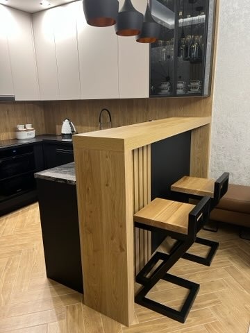 Nowoczesna wyspa kuchenna z drewnianym blatem i metalowymi hokerami na tle jasnej kuchni z czarnymi akcentami i parkietem ułożonym w jodełkę.