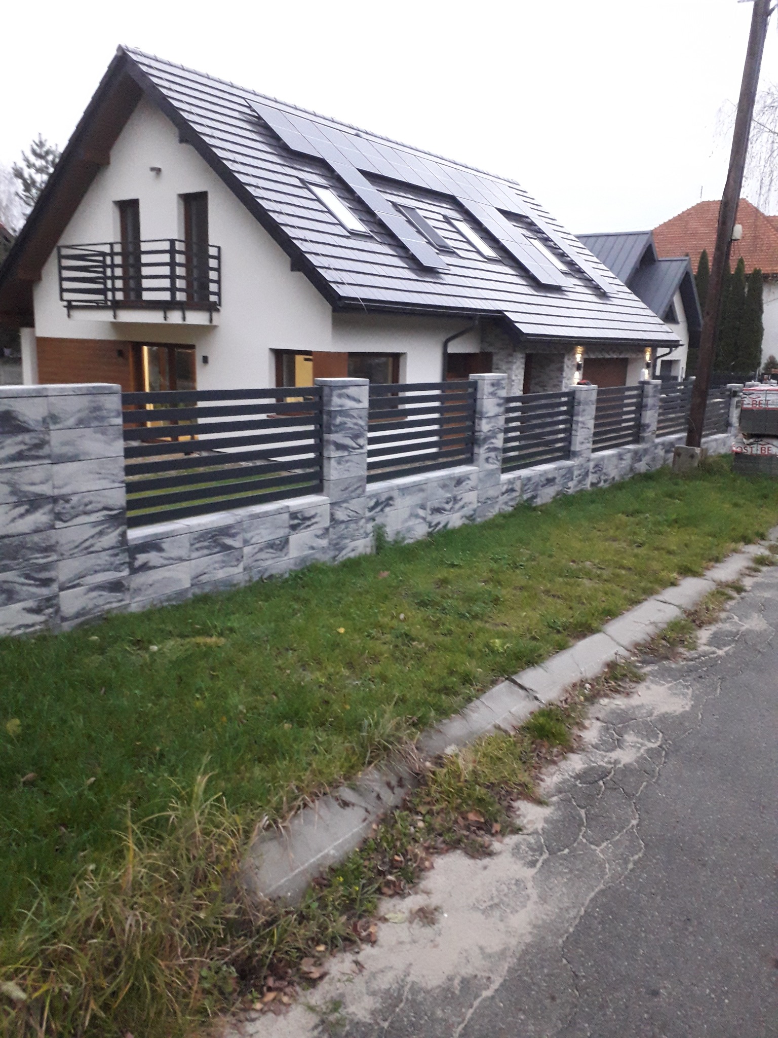 Nowoczesne ogrodzenie posesji z szarej kostki i metalowych przęseł, na tle domu z panelami słonecznymi i balkonem z czarną balustradą. Widok z poziomu ulicy.