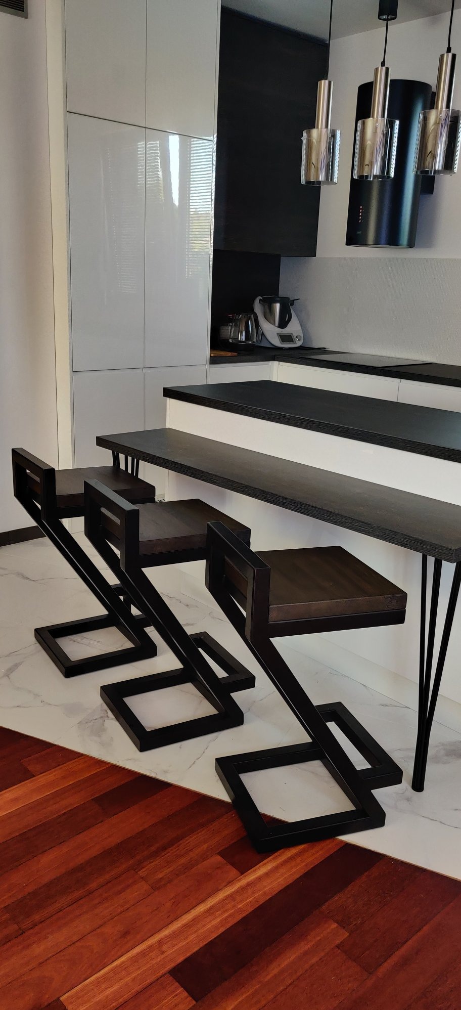 Nowoczesne, metalowe hokery barowe z ciemnego drewna i czarnych, geometrycznych podstaw, ustawione przy blacie w jasnej kuchni. Minimalistyczny design.