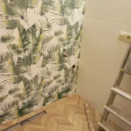 Łazienka w trakcie remontu: ułożona podłoga z paneli w jodełkę, ściana z płytek w tropikalny wzór, widoczne otwory na armaturę, drabina oparta o ścianę.
