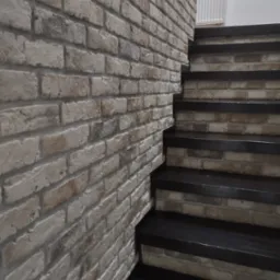 Ściana z dekoracyjnej cegły w odcieniach szarości i beżu, sąsiadująca z czarnymi schodami z drewnianymi stopniami, widoczny fragment grzejnika na białej ścianie w tle.