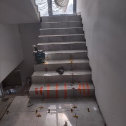 KS Remontix Krzysztof Stolc - Schody w trakcie układania płytek, widoczne poziomice, krzyżyki dystansowe, taśma malarska i wiadro z klejem. W tle okno zasłonięte folią malarską.