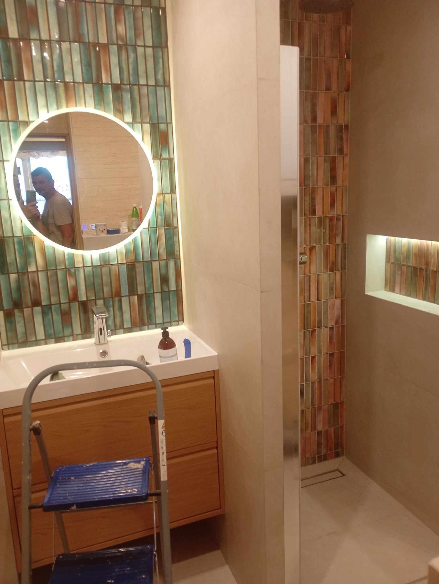 Nowoczesna łazienka z umywalką na drewnianej szafce, lustrem z podświetleniem LED i kabiną prysznicową wyłożoną mozaiką. Widoczna drabina malarska.