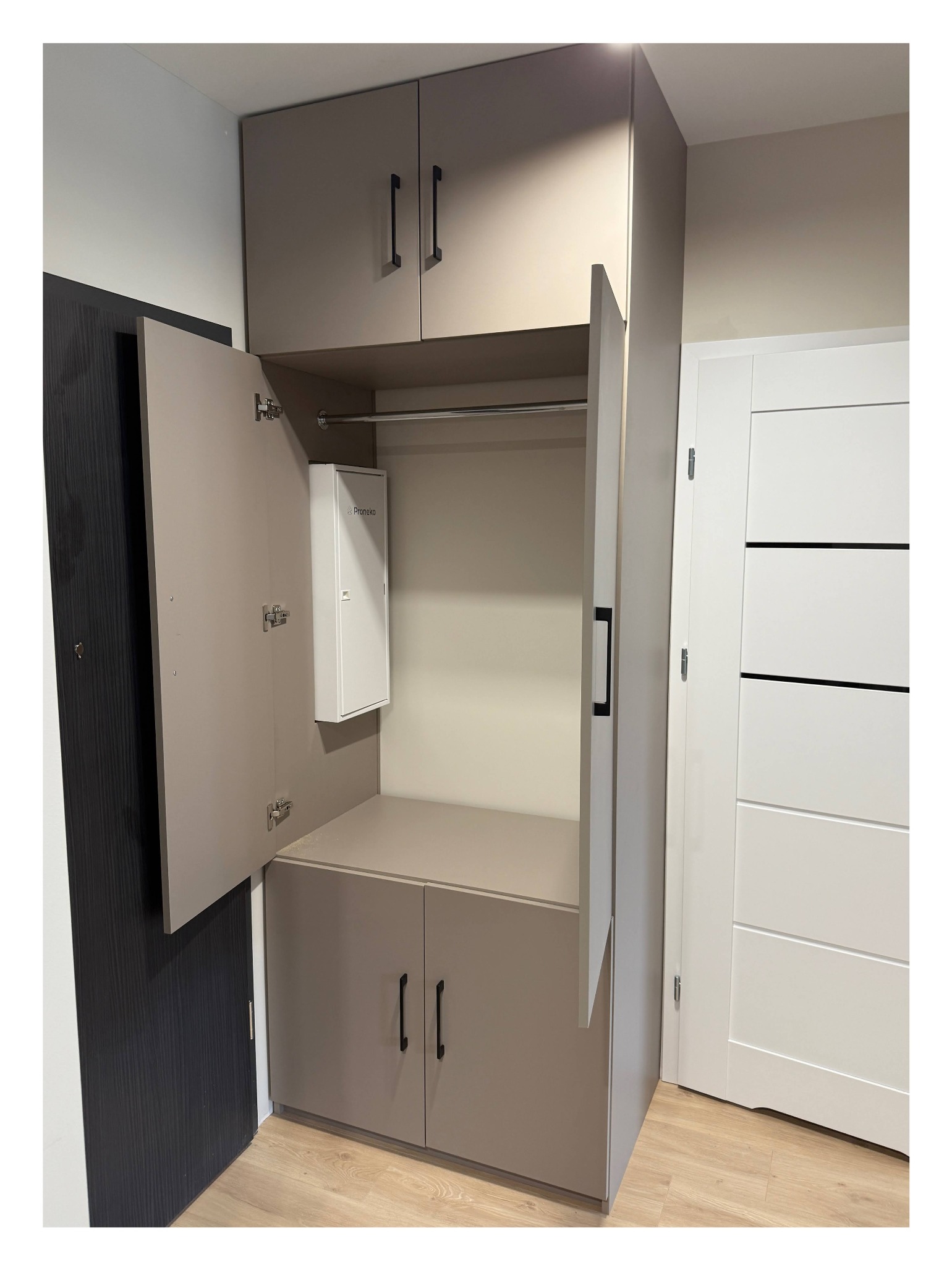 Szafa na wymiar w przedpokoju, otwarta, z widocznym wieszakiem i skrzynką Pronenko. Fronty w kolorze taupe, czarne uchwyty, drewniana podłoga.