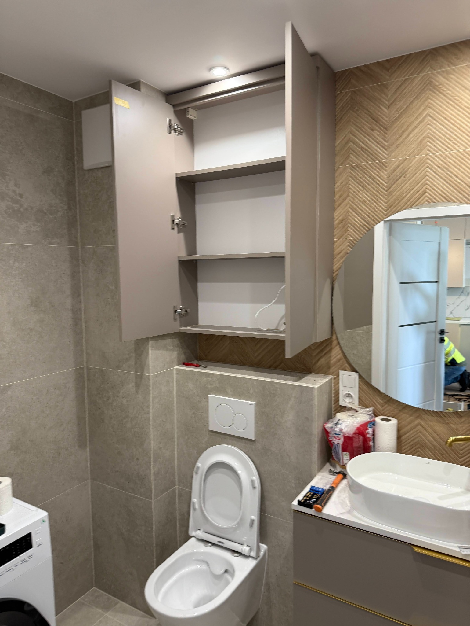 Nowoczesna łazienka z otwartą szafką wiszącą nad toaletą. Umywalka nablatowa i lustro owalne dopełniają aranżację. W tle widoczny fragment instalacji.