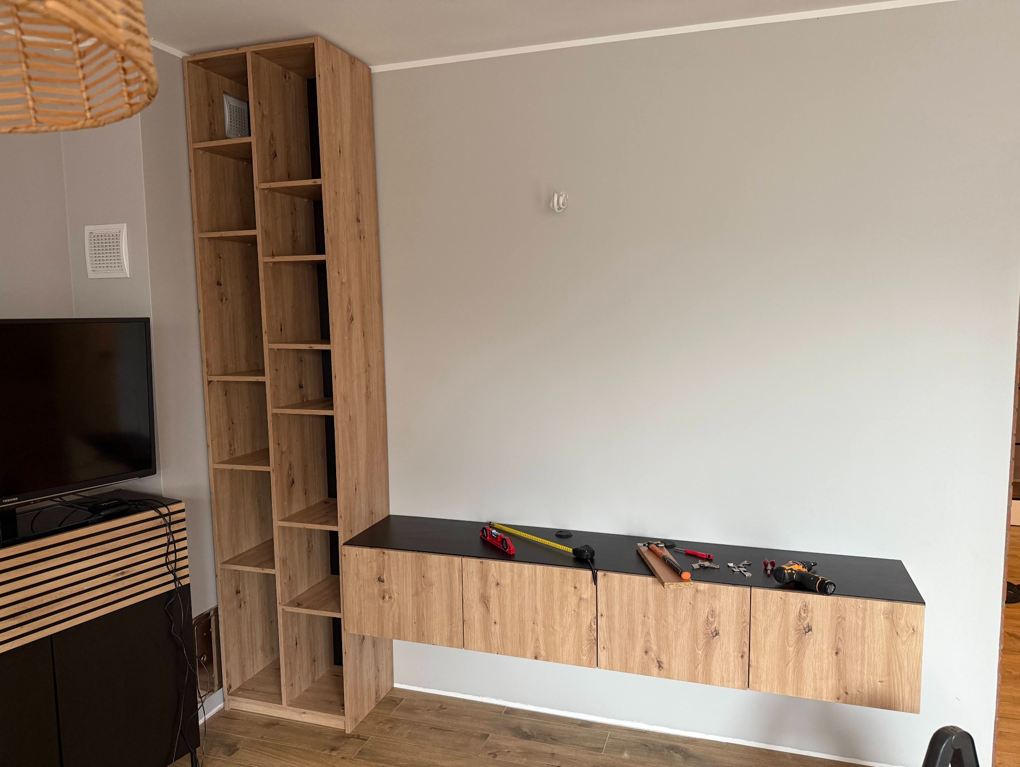 Zmontowana szafka RTV z drewnianymi frontami i czarnym blatem, narzędzia na wierzchu, wysoki regał obok. Widok na ścianę i podłogę.