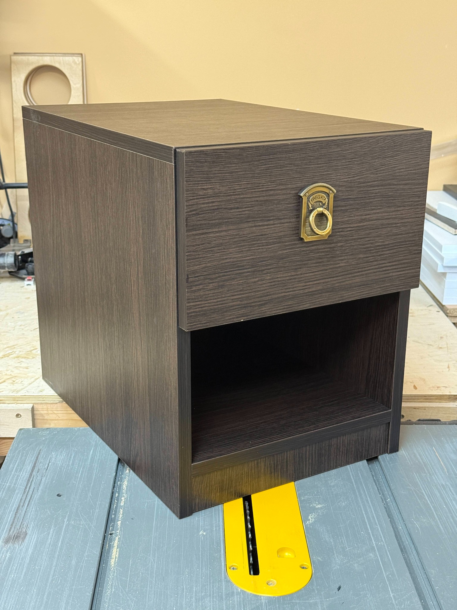 Brązowa szafka nocna z szufladą i otwartą półką, uchwyt szuflady w stylu retro, stoi na stole warsztatowym z piłą tarczową. Widoczne narzędzia stolarskie.