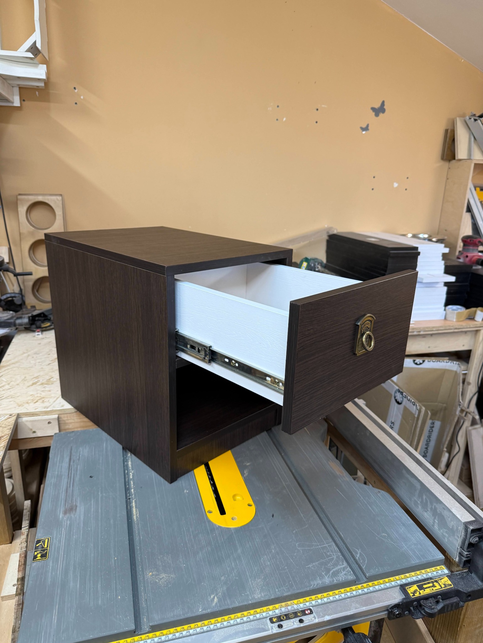 Ciemnobrązowa szafka nocna z wysuwaną, białą szufladą i ozdobnym uchwytem, ustawiona na stole warsztatowym z piłą tarczową. Widoczne narzędzia i materiały stolarskie.