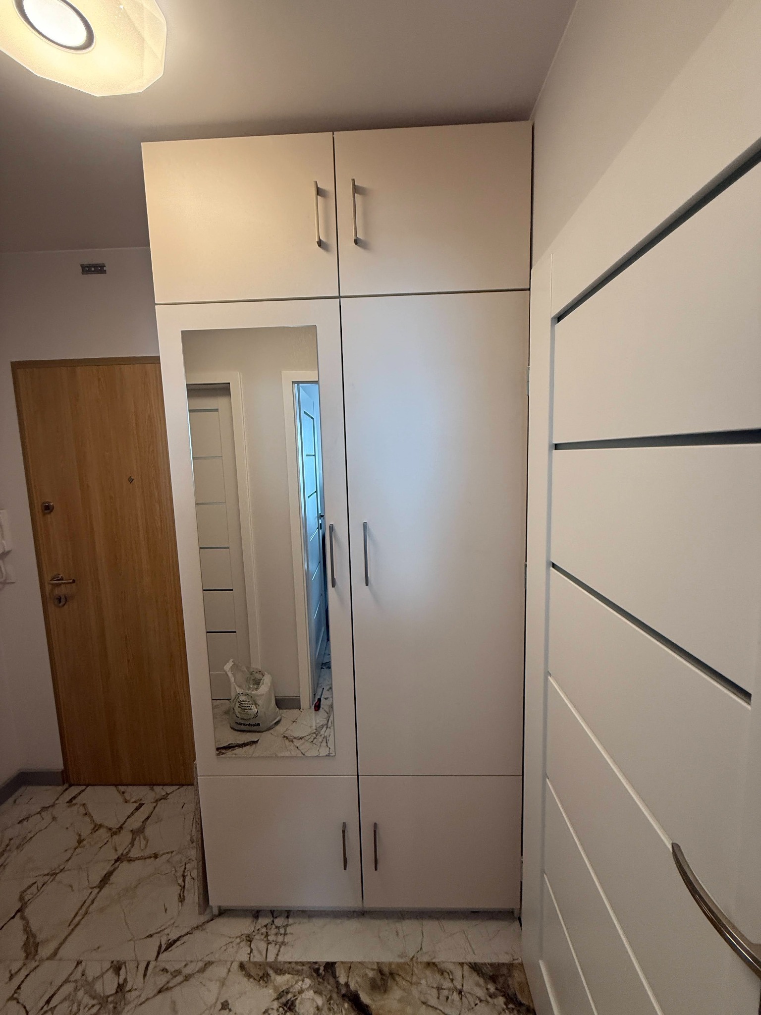 Biała szafa na wymiar z lustrem w przedpokoju w Kowalach. Widoczna podłoga imitująca marmur i drzwi wejściowe. Minimalistyczny design, pionowe uchwyty.