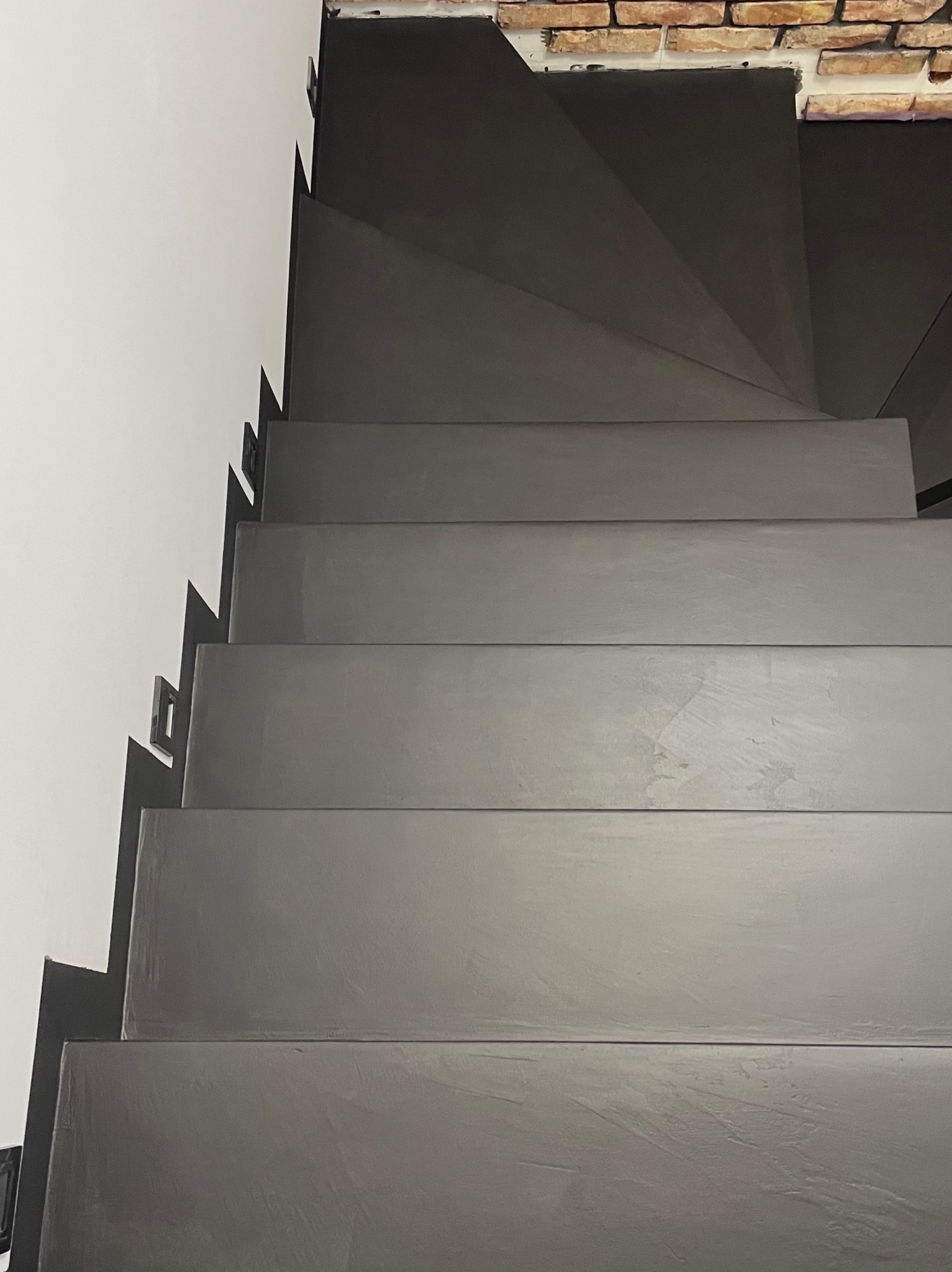 Schody spiralne wykończone szarą powłoką, widok z góry, z fragmentem białej ściany i ceglanym wykończeniem u góry.