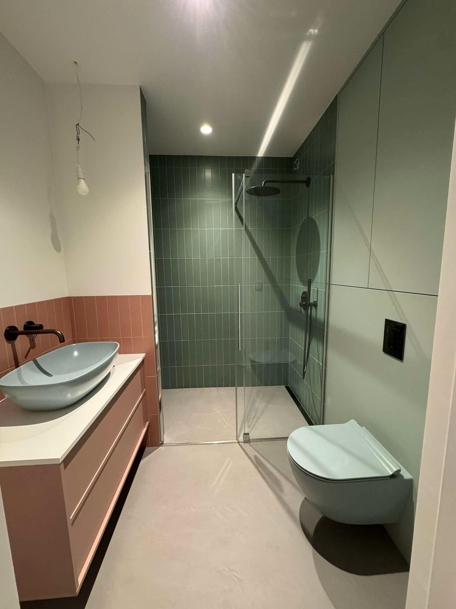 Nowoczesna łazienka w odcieniach zieleni i różu z umywalką nablatową w kształcie misy, kabiną prysznicową walk-in z zielonymi płytkami oraz podwieszanym WC w kolorze ścian, w trakcie wykańczania...