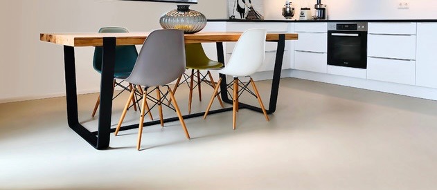 Jasna, jednolita posadzka w nowoczesnej kuchni z drewnianym stołem na metalowych nogach i krzesłami Eames, białe szafki kuchenne w tle.
