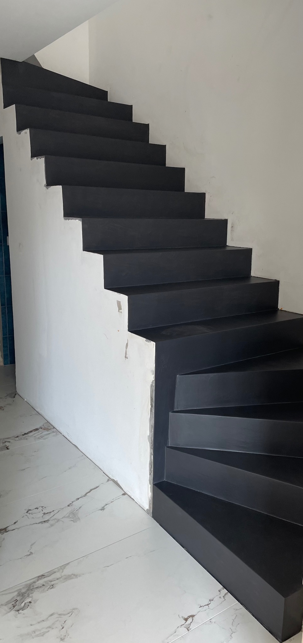 Schody wewnętrzne z czarnym wykończeniem, widoczne niedoskonałości na ścianie i marmurowe płytki podłogowe.