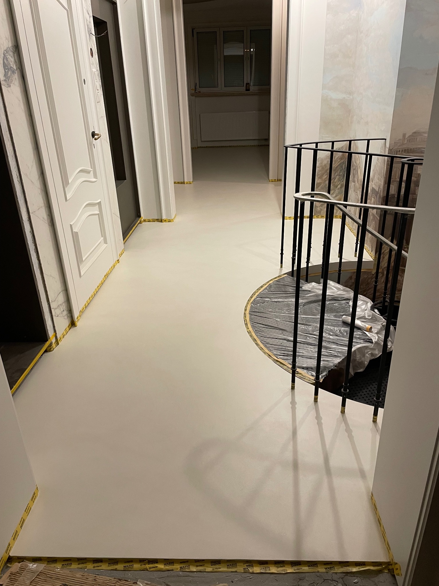 Jasna, gładka posadzka w korytarzu z zabezpieczoną taśmą malarską dolną krawędzią ścian i ościeżnic, widoczne zabezpieczone schody spiralne.