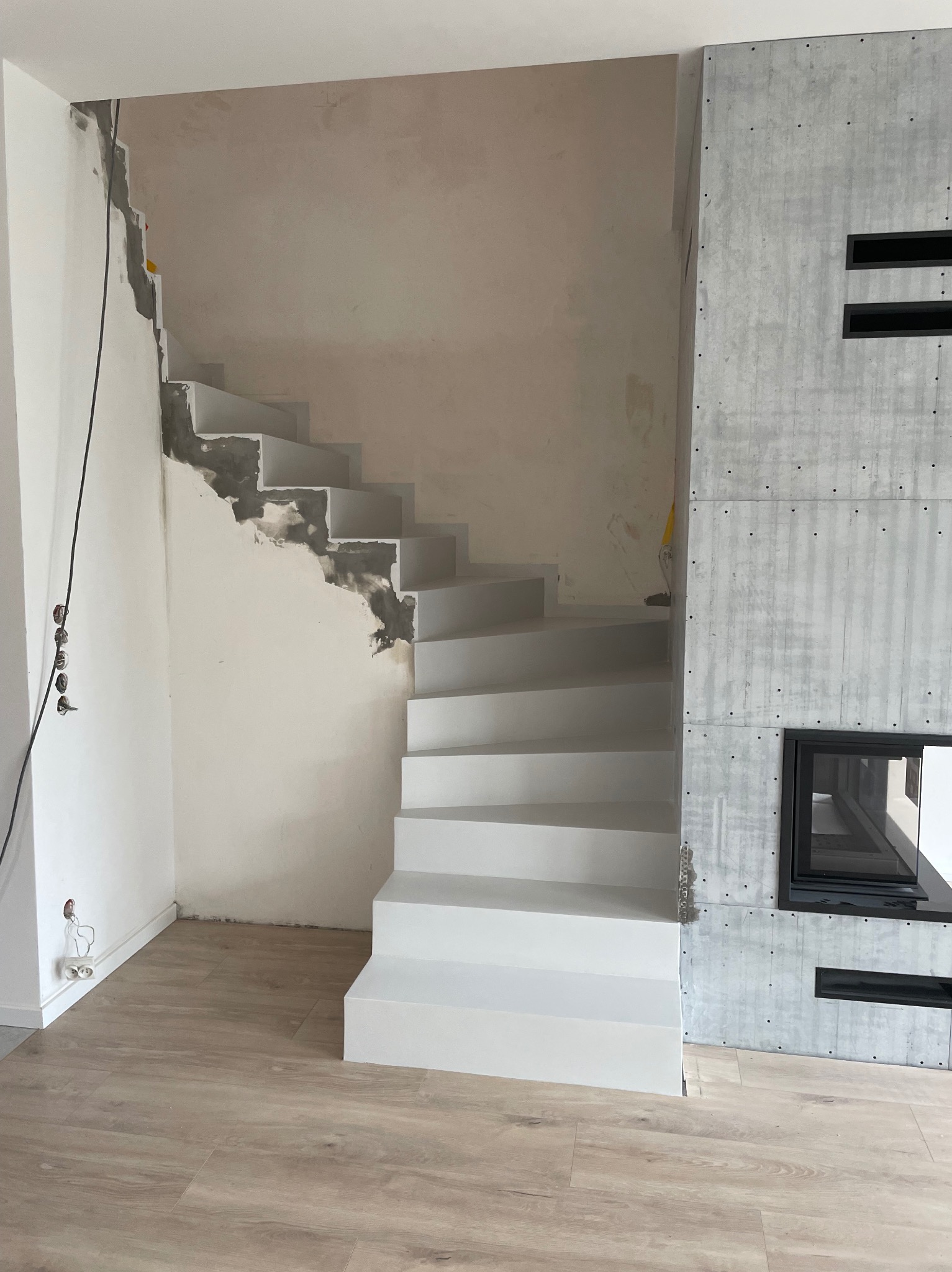 Montaż schodów z betonu architektonicznego w surowym stanie, widoczne ślady po szpachlowaniu na ścianie, obok betonowy kominek z czarnym wkładem, podłoga wyłożona panelami imitującymi drewno.