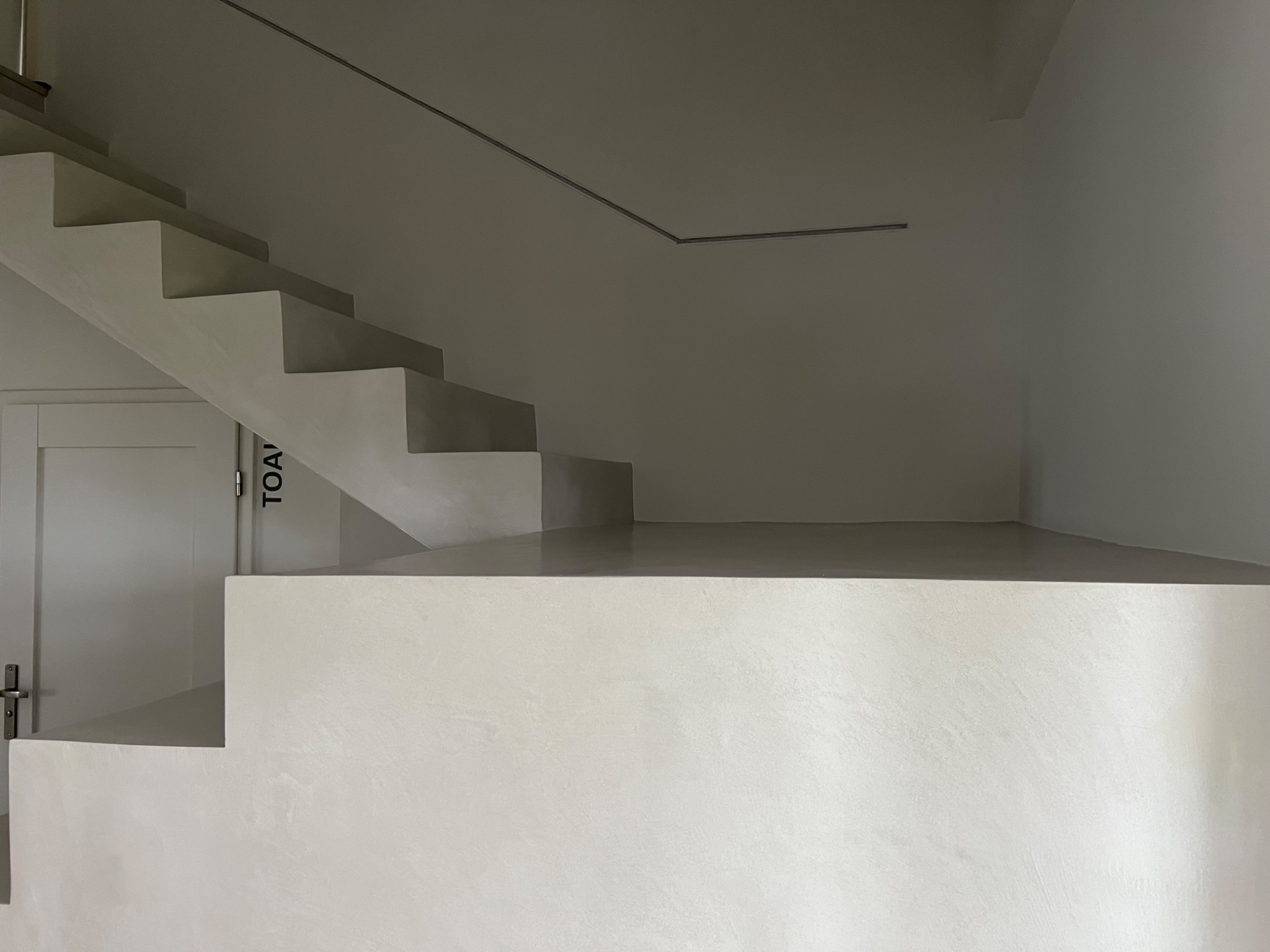 Minimalistyczne schody z mikrocementu w jasnym odcieniu szarości, prowadzące w górę, obok białe drzwi z napisem 'TOA' w pionie, widoczne metalowe wykończenie na suficie.