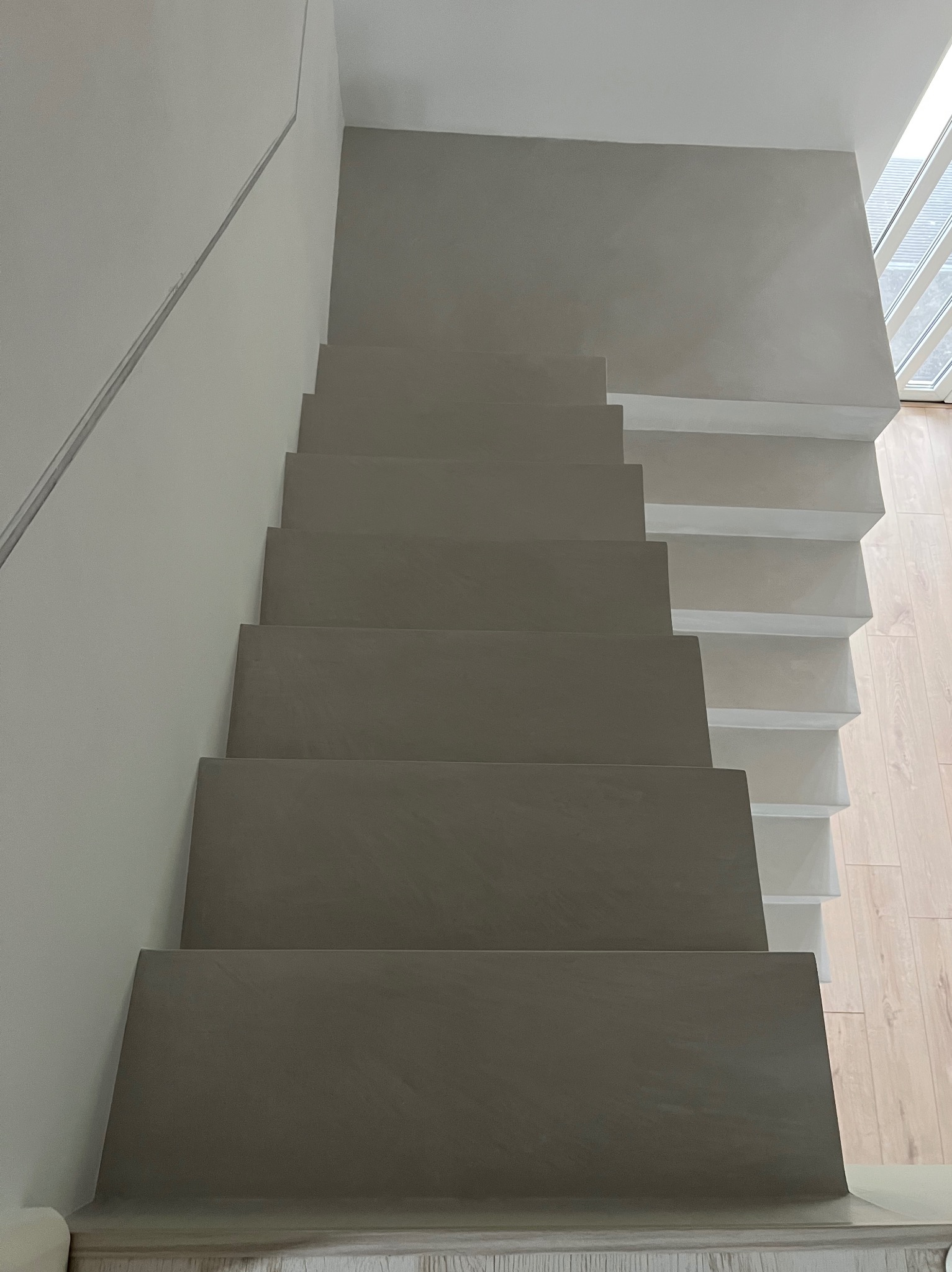 Schody pokryte szarą, gładką powłoką, widoczne z dołu, z białymi elementami konstrukcyjnymi i drewnianą podłogą w tle.