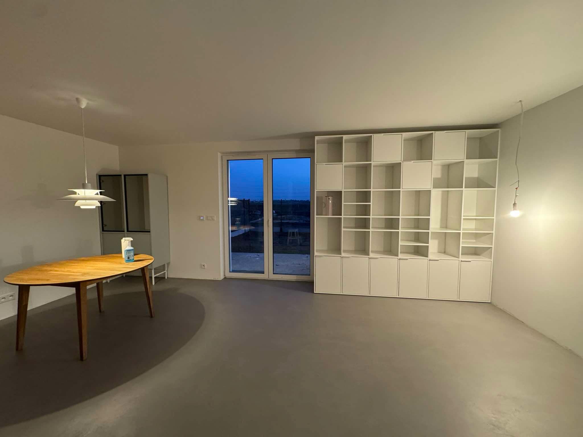 Wnętrze pokoju z szarą podłogą, drewnianym stołem z płynem do dezynfekcji, białą regałem i dużym oknem balkonowym.