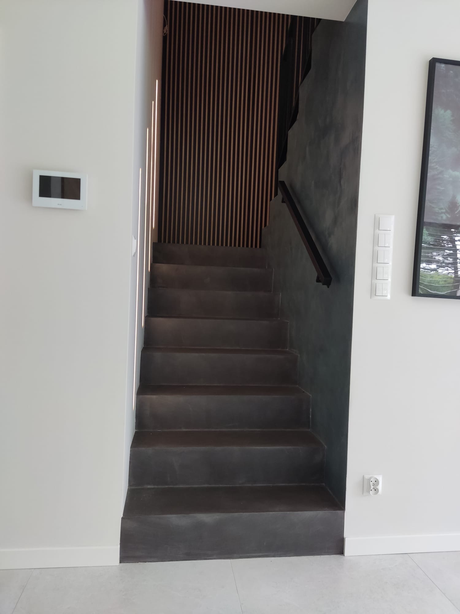 Schody w nowoczesnym domu, wykończone w ciemnym odcieniu, z drewnianą ścianą dekoracyjną i minimalistycznym oświetleniem. Widoczne elementy wykończenia ścian.