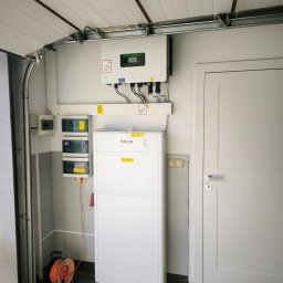 ECOAUTOMATION DAWID I WOJCIECH ZAHRT SPÓŁKA CYWILNA - Instalacja fotowoltaiczna w garażu: biały inwerter i akumulator Sofar Solar zamontowane na ścianie, widoczne zabezpieczenia elektryczne i zwinięty przedłużacz na podłodze.