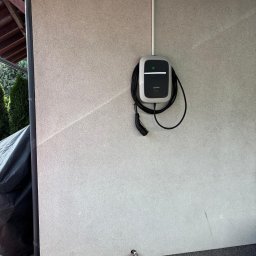ECOAUTOMATION DAWID I WOJCIECH ZAHRT SPÓŁKA CYWILNA - Zamontowana na zewnątrz budynku stacja ładowania pojazdów elektrycznych, kabel wpięty, widoczne rury instalacyjne na ścianie, fragment dachu i roślinności w tle.