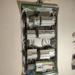 Otwarta skrzynka rozdzielcza z bezpiecznikami i okablowaniem na ścianie w trakcie instalacji elektrycznej, widoczne przewody w kolorach, listwy zaciskowe i elementy zabezpieczające.