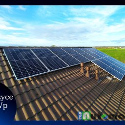 ECOAUTOMATION DAWID I WOJCIECH ZAHRT SPÓŁKA CYWILNA - Panele słoneczne zamontowane na brązowym dachu z dachówki ceramicznej, widok z góry na tle błękitnego nieba z chmurami i zielonych pól w oddali.