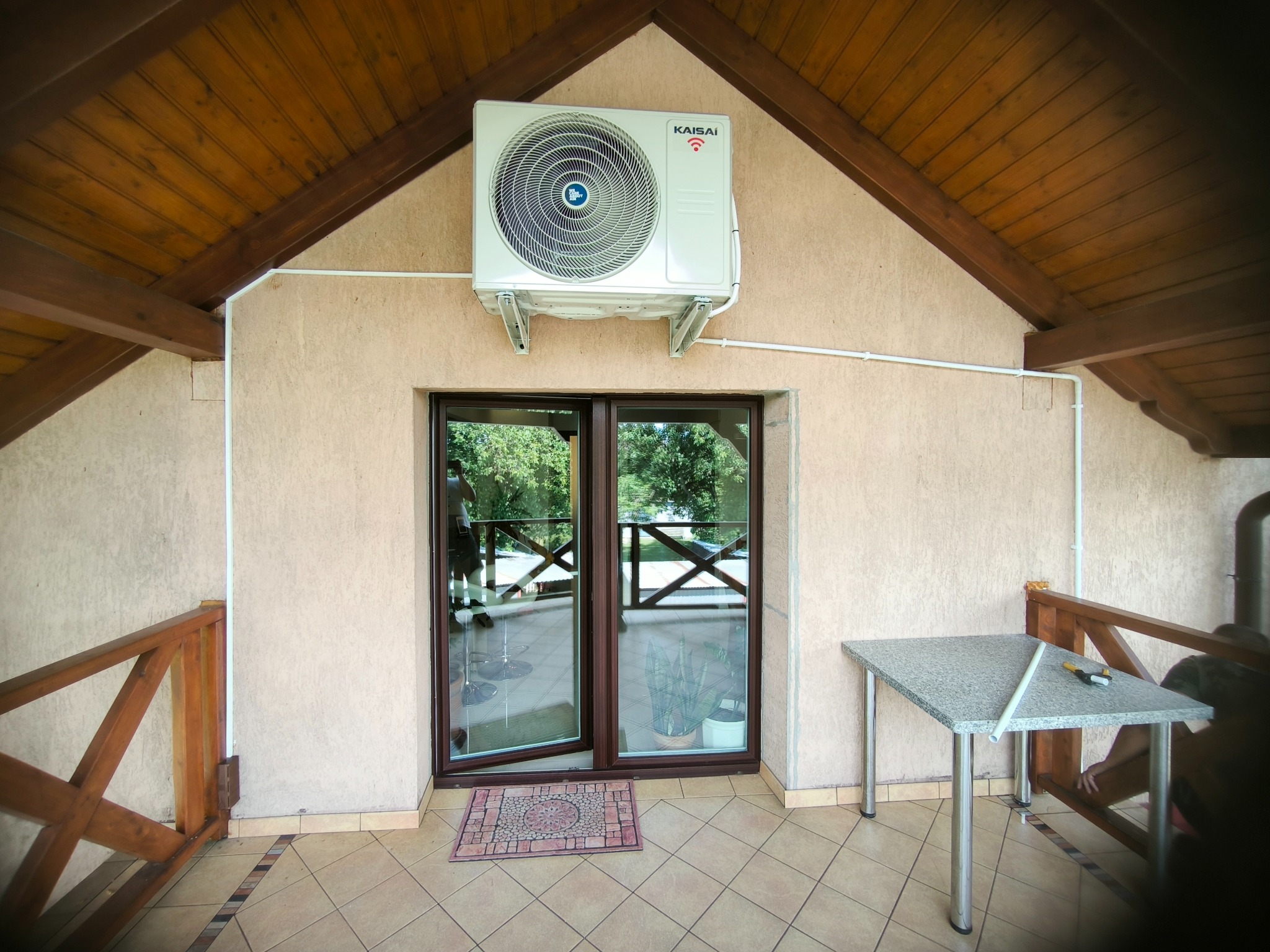Zewnętrzna jednostka klimatyzacji Kaisai zamontowana nad dwuskrzydłowymi drzwiami balkonowymi na zadaszonym tarasie z drewnianą konstrukcją dachu i kameralnym wykończeniem ścian.