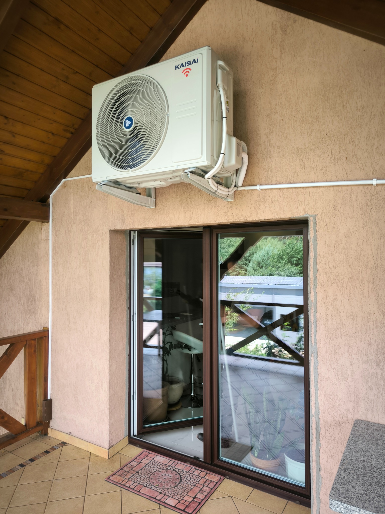 Zewnętrzna jednostka klimatyzacji marki Kaisai zamontowana na ścianie domu nad przesuwnymi drzwiami tarasowymi, widoczne białe przewody instalacyjne i drewniana konstrukcja dachu.