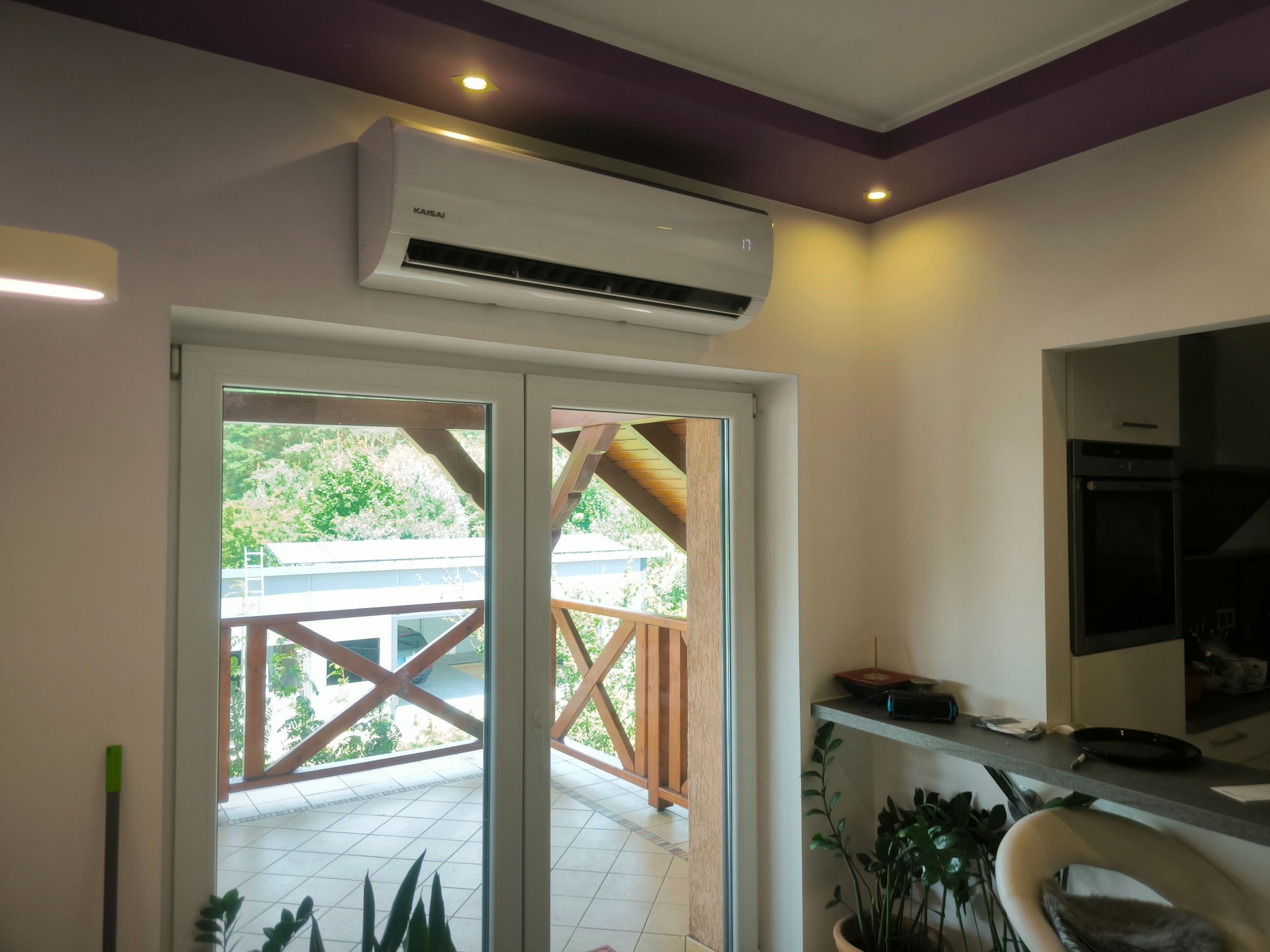 Biała klimatyzacja marki Kaisai zamontowana nad przeszklonym wyjściem na balkon, widok na drewnianą balustradę i zieleń za nią, fioletowy pas na suficie z oświetleniem punktowym.