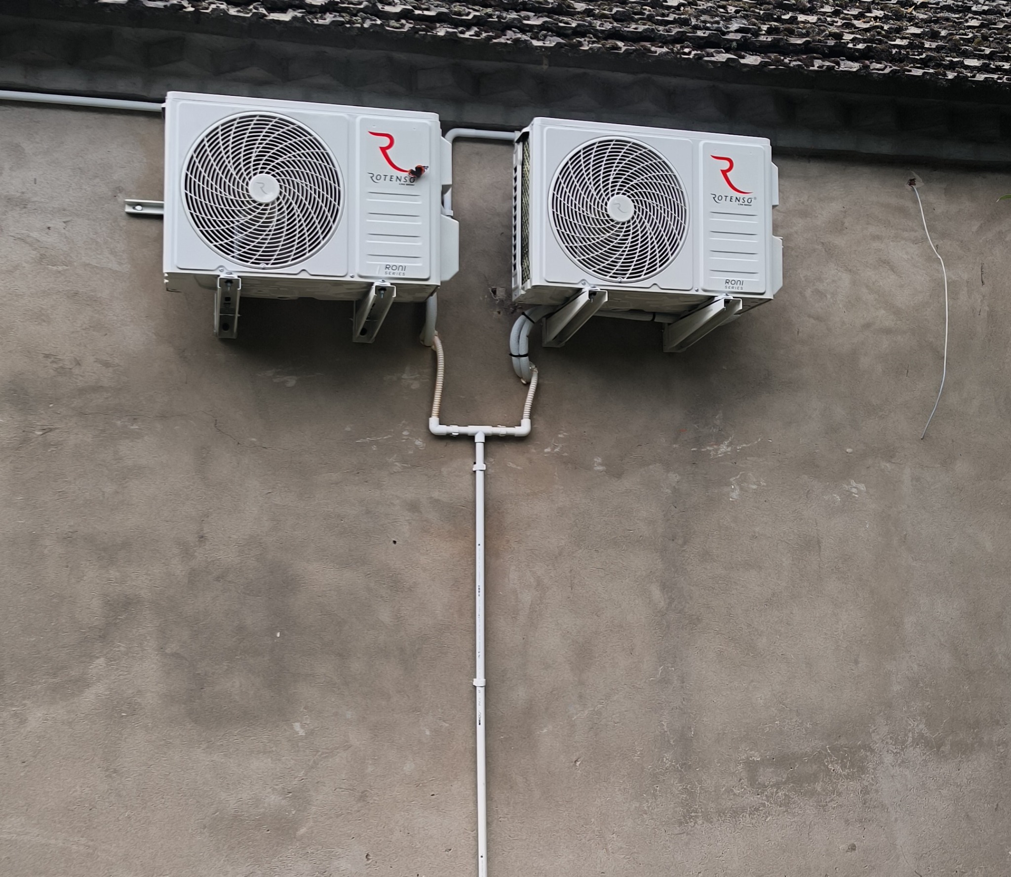 Dwa białe klimatyzatory marki Rotenso zamontowane na szarej, teksturowanej ścianie zewnętrznej budynku, z widocznymi rurami instalacyjnymi.