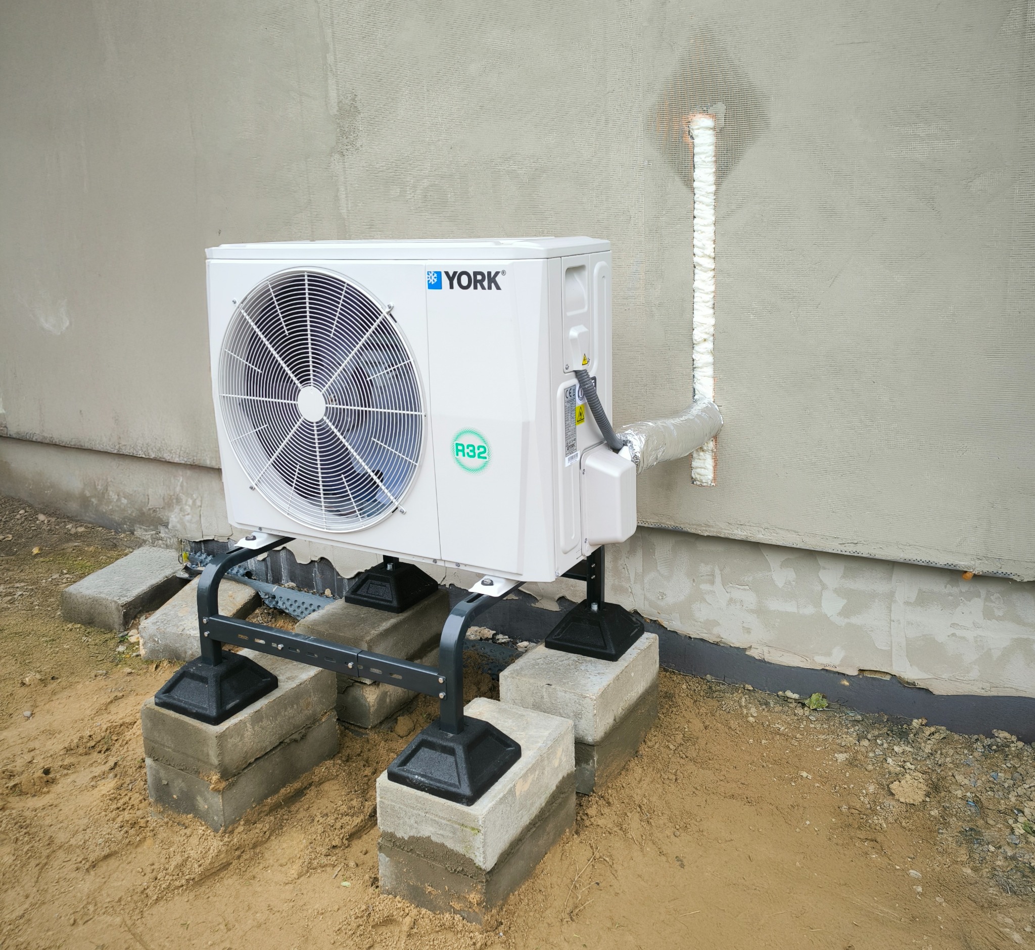 Biała jednostka zewnętrzna klimatyzacji marki York, zamontowana na betonowych bloczkach na gruncie, z widocznymi rurami zabezpieczonymi pianką montażową przy ścianie budynku w trakcie wykończenia.