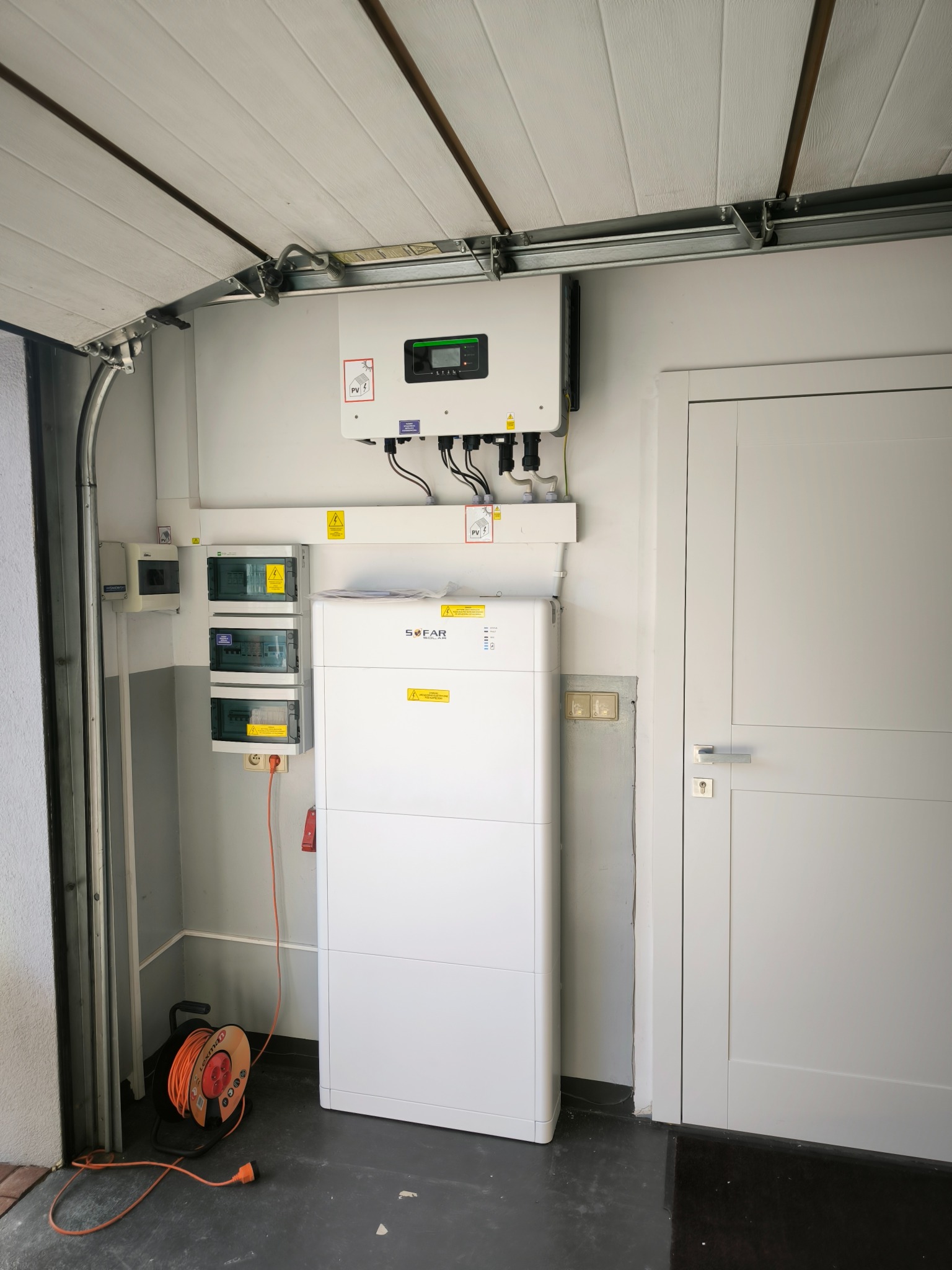 Instalacja fotowoltaiczna w garażu: biały inwerter i akumulator Sofar Solar zamontowane na ścianie, widoczne zabezpieczenia elektryczne i zwinięty przedłużacz na podłodze.