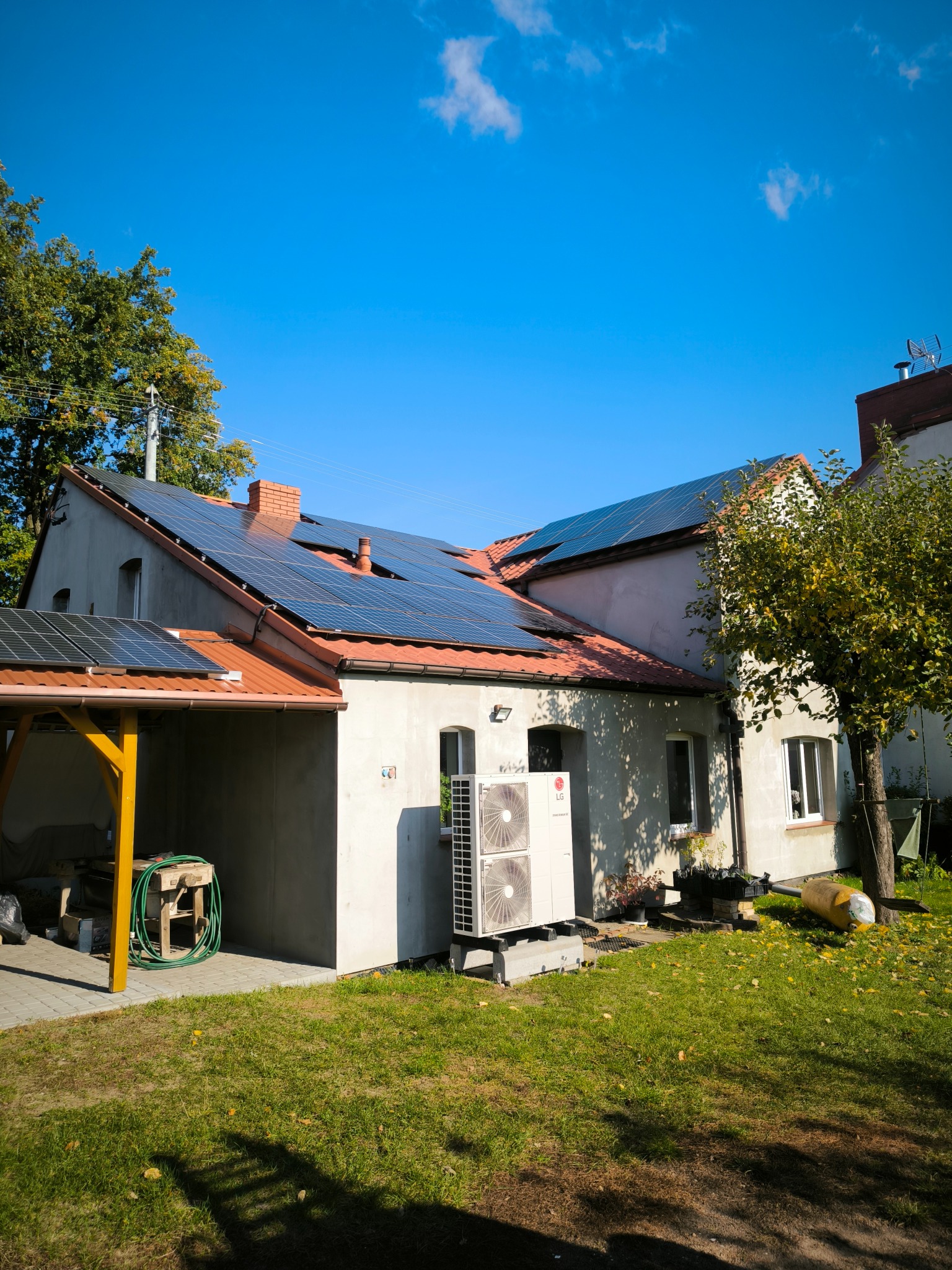 Dom jednorodzinny z zainstalowanymi panelami fotowoltaicznymi na dachu i pompą ciepła LG na zewnątrz, widok z ogrodu w słoneczny dzień.