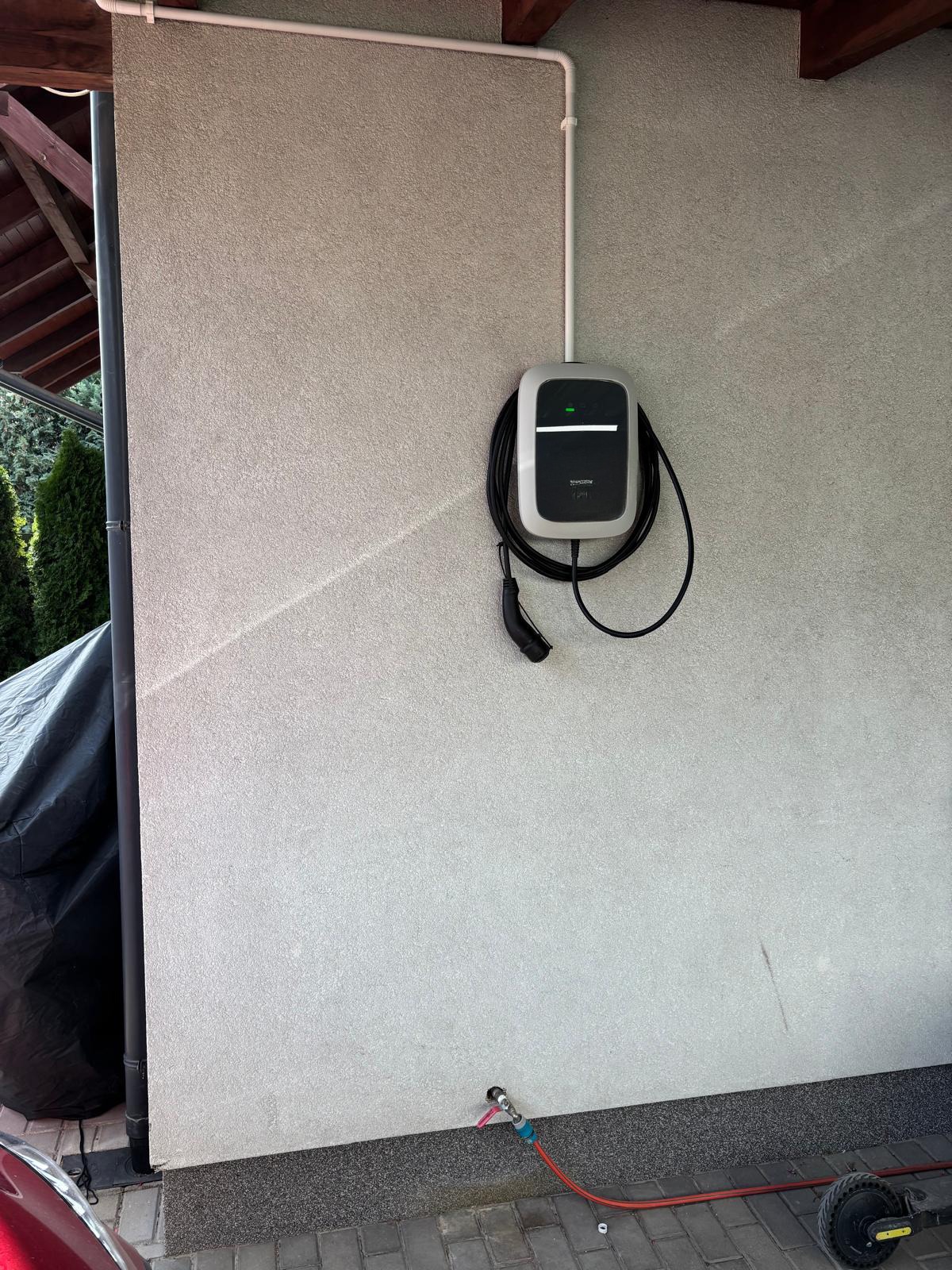Zamontowana na zewnątrz budynku stacja ładowania pojazdów elektrycznych, kabel wpięty, widoczne rury instalacyjne na ścianie, fragment dachu i roślinności w tle.