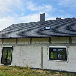 Przedsiębiorstwo Produkcyjno-Handlowo-Usługowe Prime Home Daniel Paździora - Dom w trakcie budowy z czarnymi oknami i dachem z dachówki. W oknach widoczne odbicia otoczenia. Budynek z białego bloczka, otoczony zielenią i błękitnym niebem.