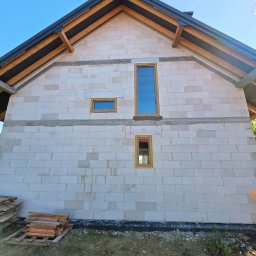 Przedsiębiorstwo Produkcyjno-Handlowo-Usługowe Prime Home Daniel Paździora - Ściana domu w trakcie budowy z zamontowanymi oknami o drewnianych ramach. Widoczne bale dachowe i niebieskie niebo. Obok leżą drewniane deski.