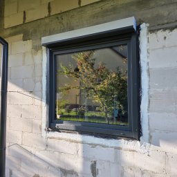 Przedsiębiorstwo Produkcyjno-Handlowo-Usługowe Prime Home Daniel Paździora - Szare okno z roletą zewnętrzną w surowej ścianie z betonu komórkowego, widoczne ślady po montażu, odbicie drzewa w szybie, rura spustowa obok.