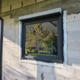 Szare okno z roletą zewnętrzną w surowej ścianie z betonu komórkowego, widoczne ślady po montażu, odbicie drzewa w szybie, rura spustowa obok.