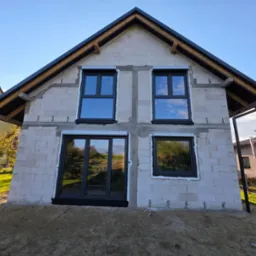 Surowy budynek z zamontowanymi oknami w grafitowej ramie, widoczne refleksy nieba w szybach. Dom w trakcie budowy, ściany z białego bloczka.