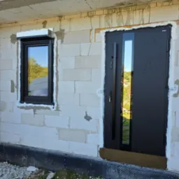 Ciemne drzwi antywłamaniowe z szybą i okno z roletą zewnętrzną w surowym stanie budynku z białego bloczka, widok z zewnątrz.